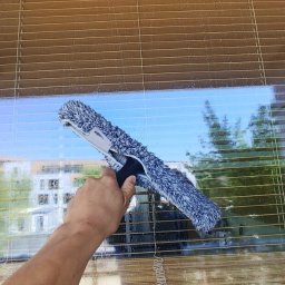 Molly - Mycie okna z żaluzjami za pomocą mopa z mikrofibry. Widoczne krople wody na szybie i odbicie budynku na zewnątrz. Żółta rama okienna.