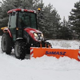 Molly - Czerwony traktor Samasz T503 z pomarańczowym pługiem odśnieżającym, usuwający śnieg z drogi w zimowy dzień. W tle ośnieżone drzewa i krzewy.