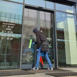 Molly - Mężczyzna w niebieskich rękawicach myje okno budynku za pomocą ściągaczki. Widoczne godziny otwarcia salonu sprzedaży i serwisu.