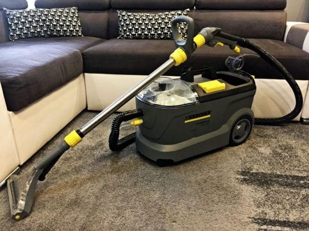 Profesjonalny odkurzacz piorący Kärcher na dywanie w salonie, gotowy do użycia. W tle widoczny narożnik z poduszkami w geometryczne wzory.
