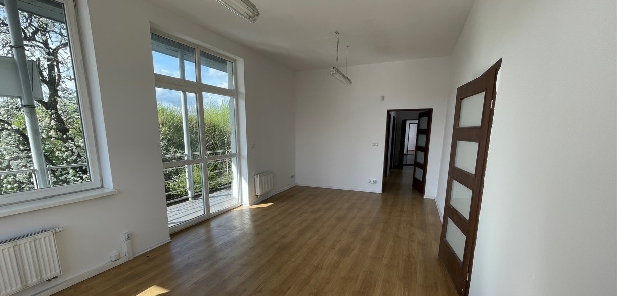 Puste, jasne biuro z parkietem, dużymi oknami z widokiem na zieleń i otwartymi drzwiami. Świeżo odmalowane ściany i minimalistyczne oświetlenie.