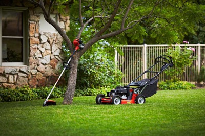 Czerwona kosiarka spalinowa i podkaszarka oparta o drzewo na tle zielonego trawnika i kamiennego domu. Sprzęt ogrodniczy po koszeniu trawy.