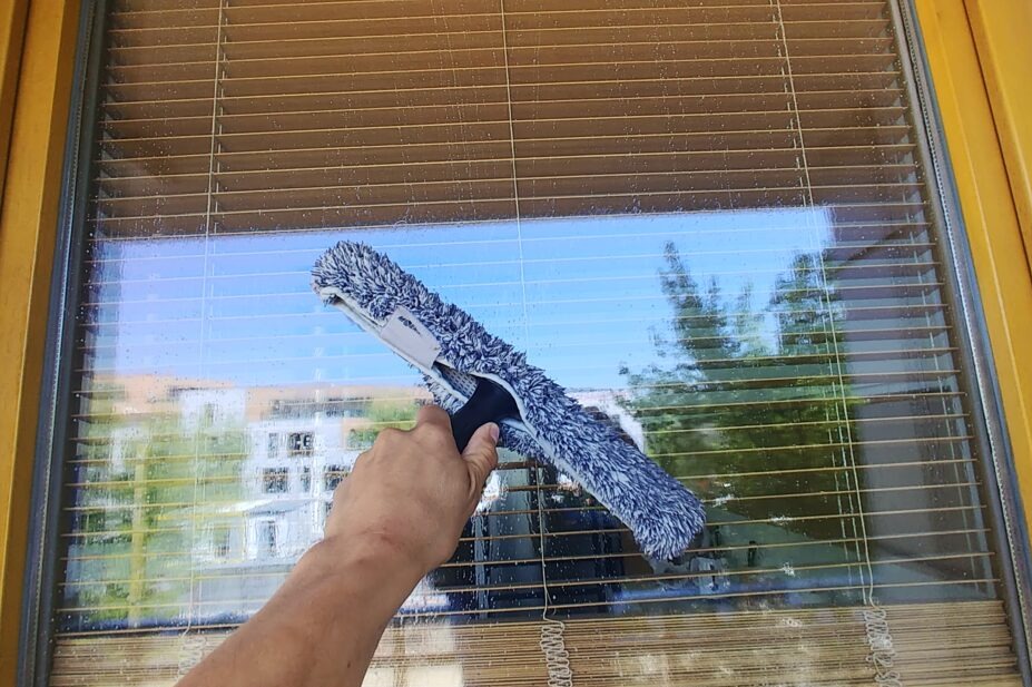 Mycie okna z żaluzjami za pomocą mopa z mikrofibry. Widoczne krople wody na szybie i odbicie budynku na zewnątrz. Żółta rama okienna.