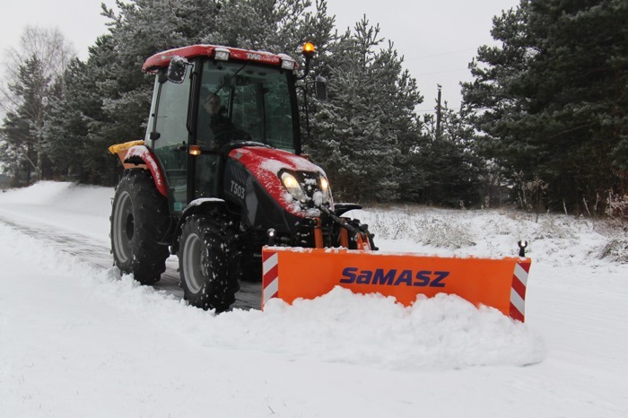 Czerwony traktor Samasz T503 z pomarańczowym pługiem odśnieżającym, usuwający śnieg z drogi w zimowy dzień. W tle ośnieżone drzewa i krzewy.