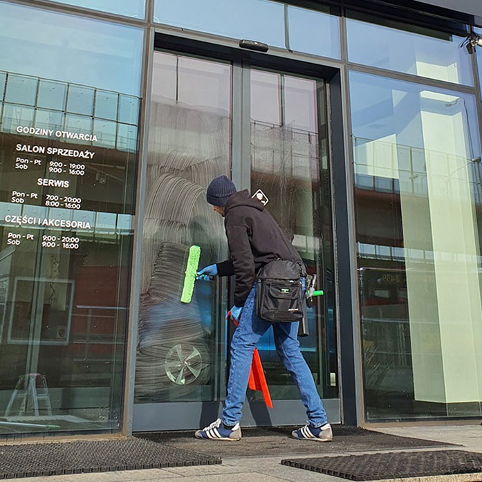 Mężczyzna w niebieskich rękawicach myje okno budynku za pomocą ściągaczki. Widoczne godziny otwarcia salonu sprzedaży i serwisu.