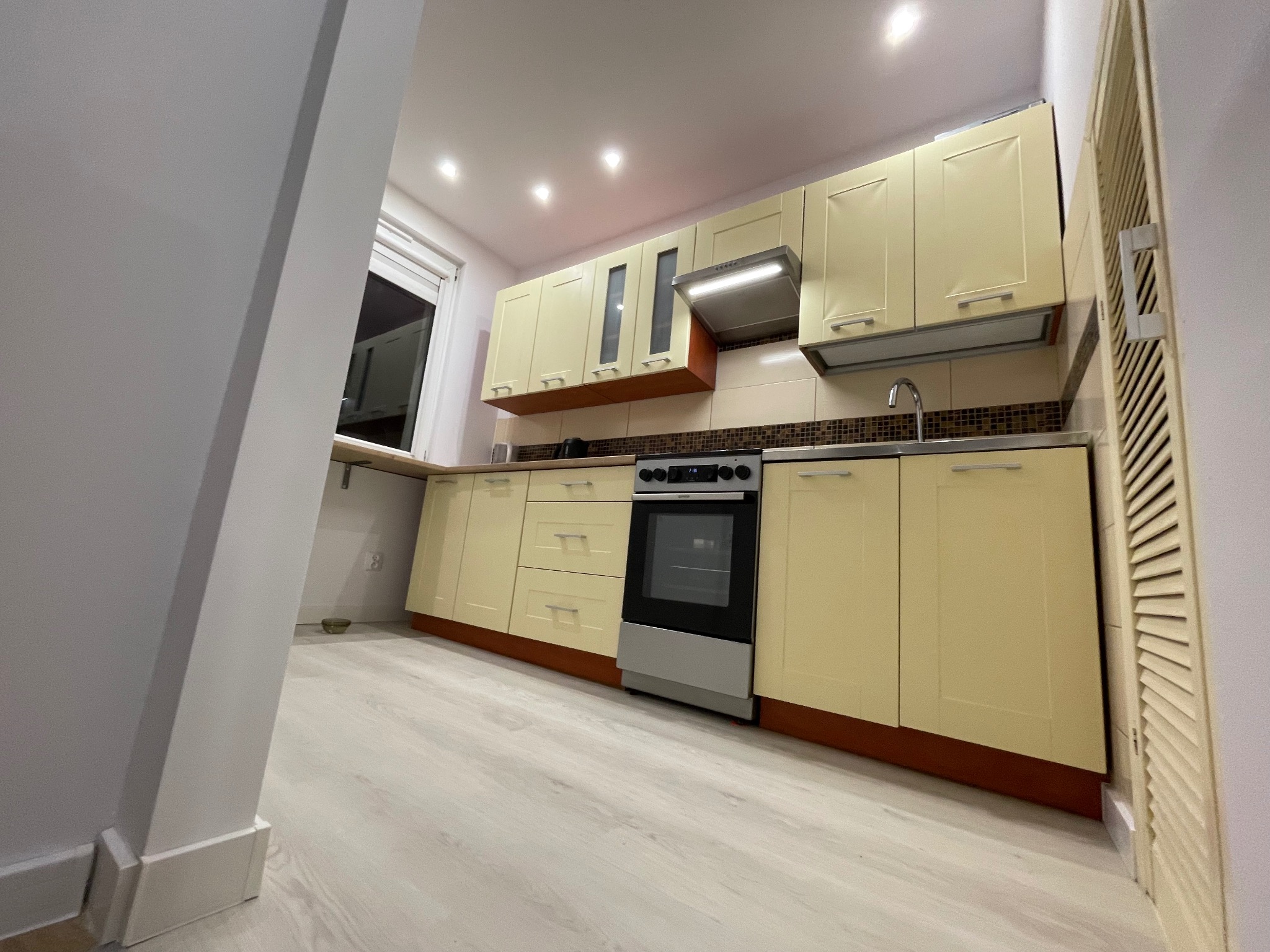 Jasna kuchnia z beżowymi szafkami i drewnianym blatem, widok z perspektywy podłogi, z widoczną kuchenką i okapem. Uwagę przykuwa nietypowe ułożenie płytek nad blatem.