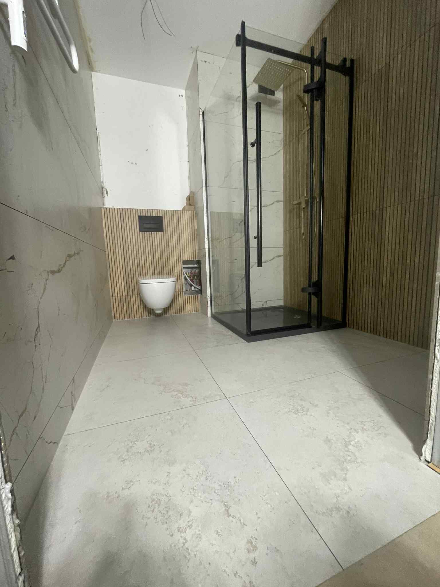 Nowoczesna łazienka w trakcie wykańczania: kabina prysznicowa z czarną ramą, podwieszana toaleta i marmurowe płytki ścienne z drewnianymi lamelami.