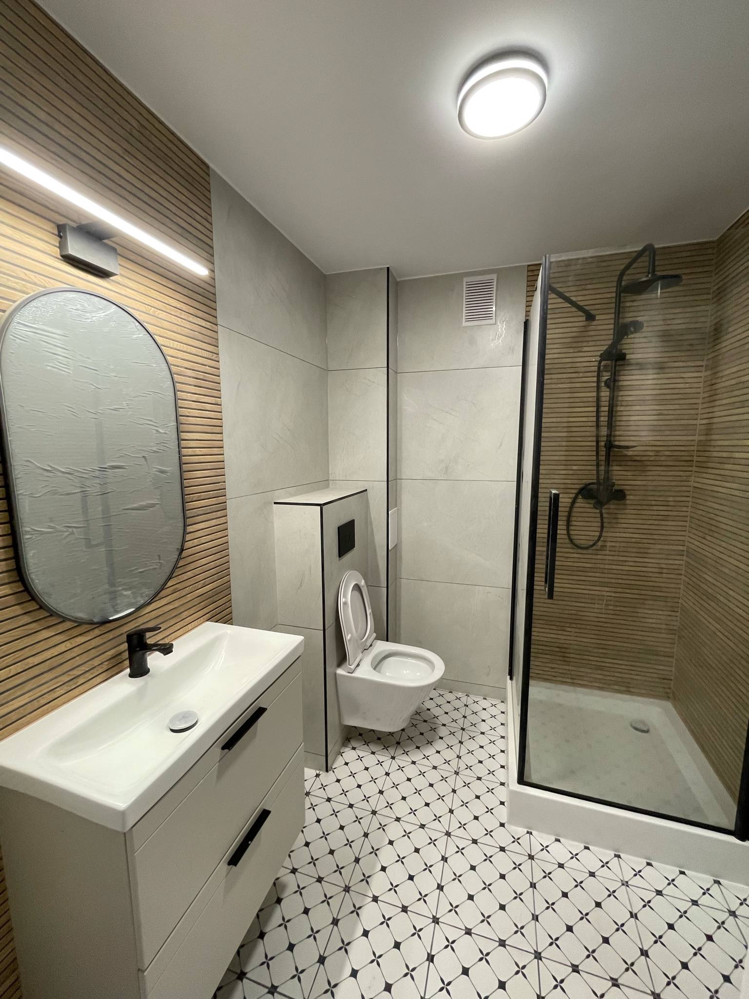 Nowoczesna łazienka po remoncie: kabina prysznicowa z czarnymi ramami, umywalka z szafką, toaleta w zabudowie i wzorzysta podłoga. Drewnopodobne płytki na ścianie.