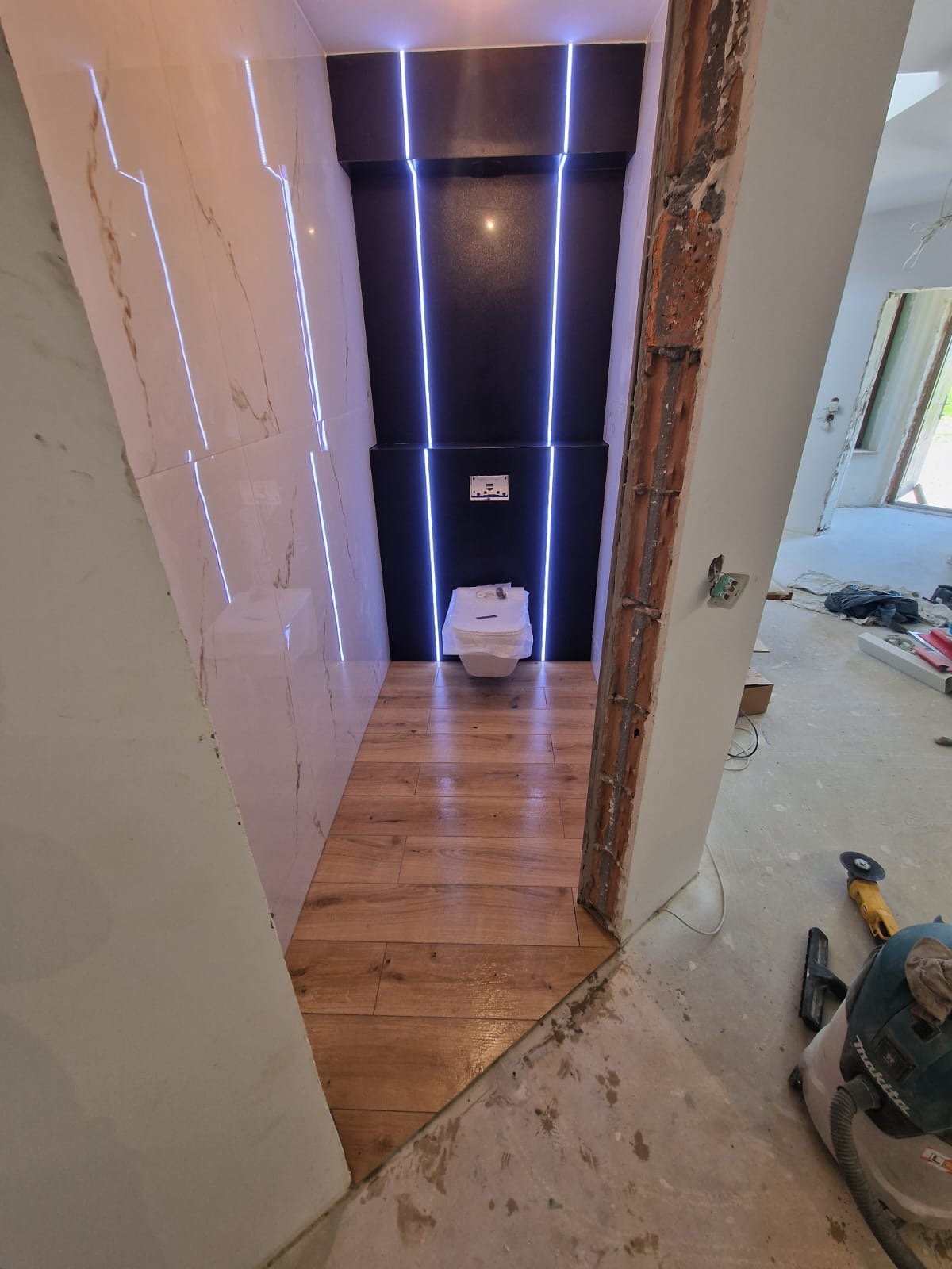 Nowoczesna łazienka w trakcie wykończenia: drewnopodobna podłoga, marmurowe płytki, czarna ściana z oświetleniem LED. Widoczne narzędzia i surowy stan wykończenia.