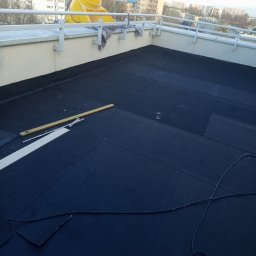 Sanmi - Czarna papa na dachu budynku z żółtym wiadrem i poziomicą. Widoczne narzędzia i fragmenty materiałów budowlanych. W tle zabudowa miejska.