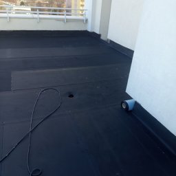 Sanmi - Wykończony dach płaski pokryty czarną membraną bitumiczną, widoczny fragment ściany i rolka materiału izolacyjnego. W tle zabudowa miejska.