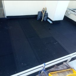 Sanmi - Wykończony dach pokryty czarną papą, widoczne narzędzia i materiały budowlane. Ujęcie z góry, z widoczną barierką balkonu.