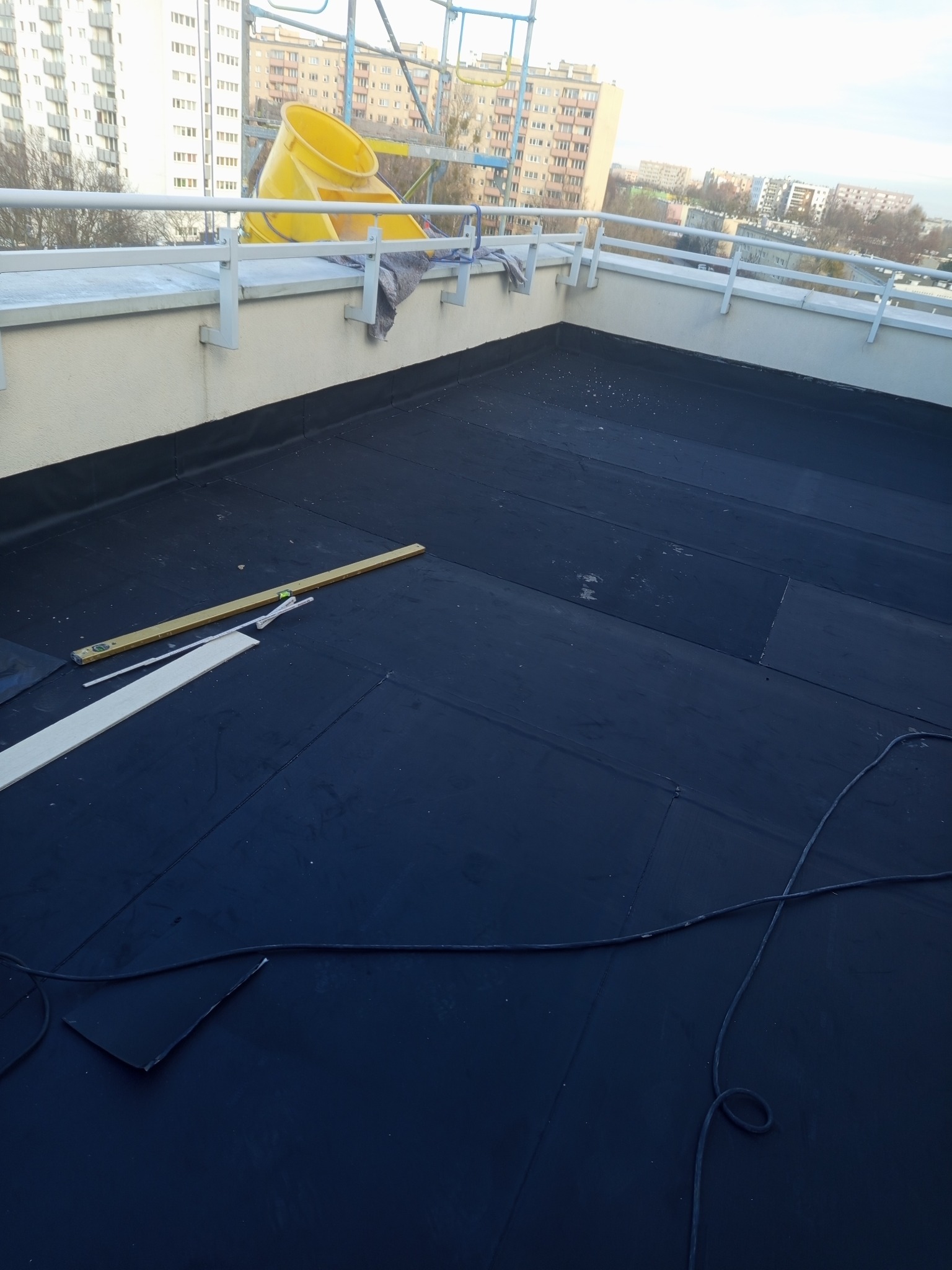 Czarna papa na dachu budynku z żółtym wiadrem i poziomicą. Widoczne narzędzia i fragmenty materiałów budowlanych. W tle zabudowa miejska.