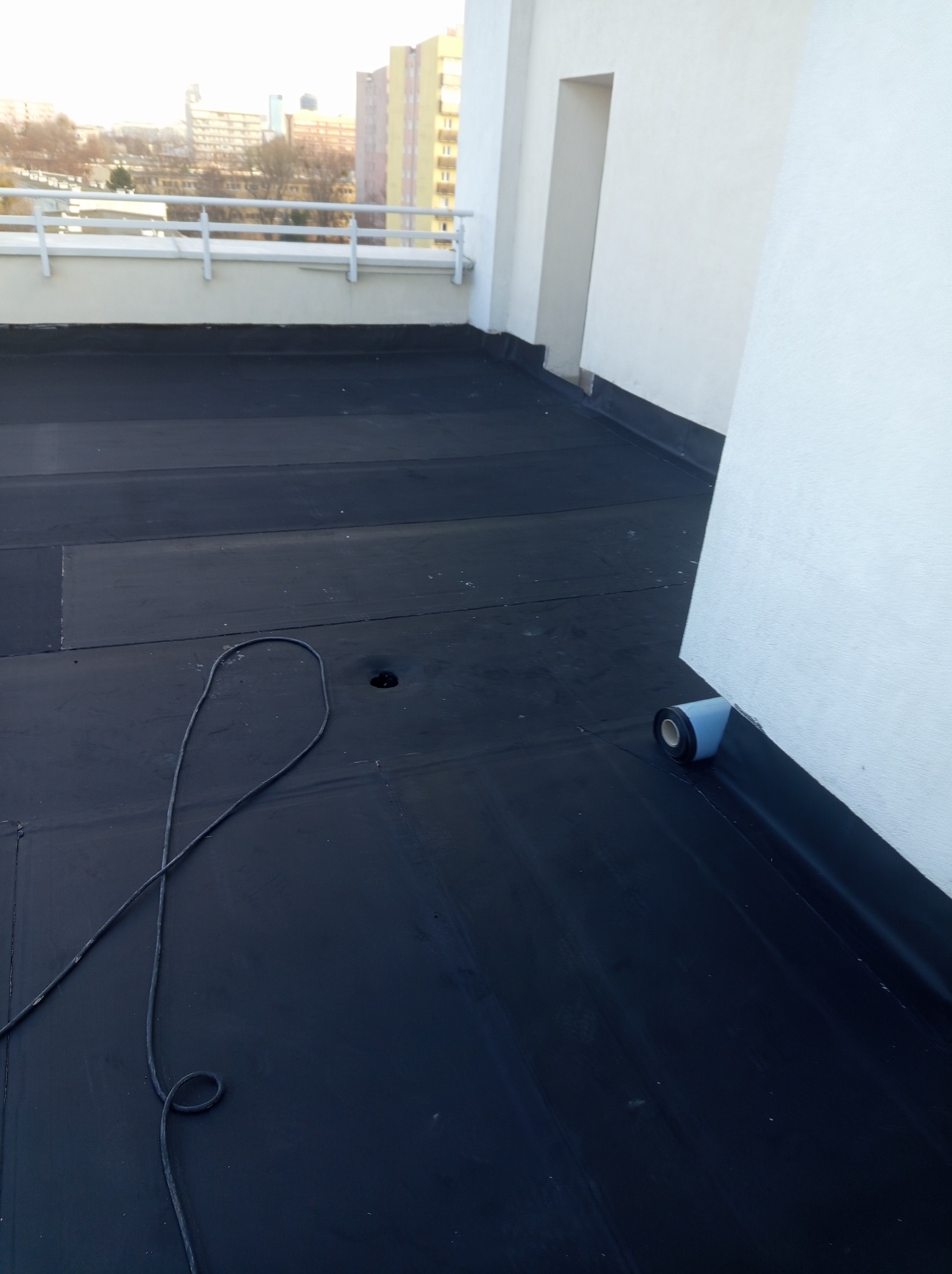 Wykończony dach płaski pokryty czarną membraną bitumiczną, widoczny fragment ściany i rolka materiału izolacyjnego. W tle zabudowa miejska.