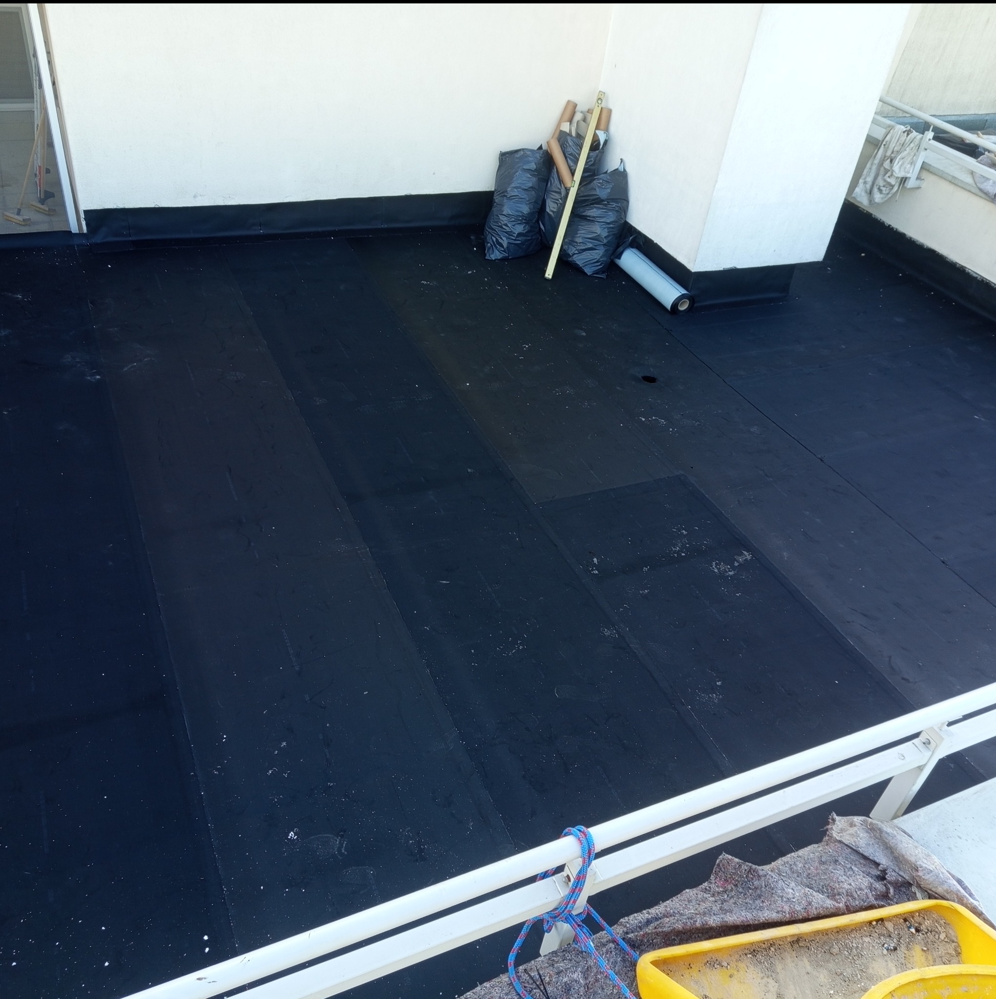 Wykończony dach pokryty czarną papą, widoczne narzędzia i materiały budowlane. Ujęcie z góry, z widoczną barierką balkonu.