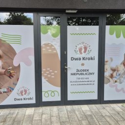 Spójnia Studio Dariusz Kawalec - Okna żłobka 'Dwa Kroki' z naklejkami prezentującymi logo, dane kontaktowe i wizerunki dzieci, tworzące przyjazną i kolorową identyfikację wizualną placówki.