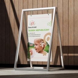 Spójnia Studio Dariusz Kawalec - Stojak reklamowy z plakatem żłobka 'Dwa Kroki' na tle drewnianej ściany. Plakat zawiera logo, dane kontaktowe i zdjęcie uśmiechniętego dziecka z kwiatem we włosach.