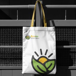 Spójnia Studio Dariusz Kawalec - Biała torba z żółtymi uchwytami, wisząca na metalowym ogrodzeniu. Na torbie logo 'Suneeco' i grafika przedstawiająca słońce w liściach. Tło rozmyte, czarno-białe.