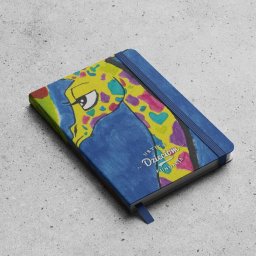 Spójnia Studio Dariusz Kawalec - Niebieski notatnik z rysunkiem żyrafy w stylu dziecięcym, leżący na szarym tle. Na okładce widoczny napis 'Urtica Dzieciom Fundacja'.