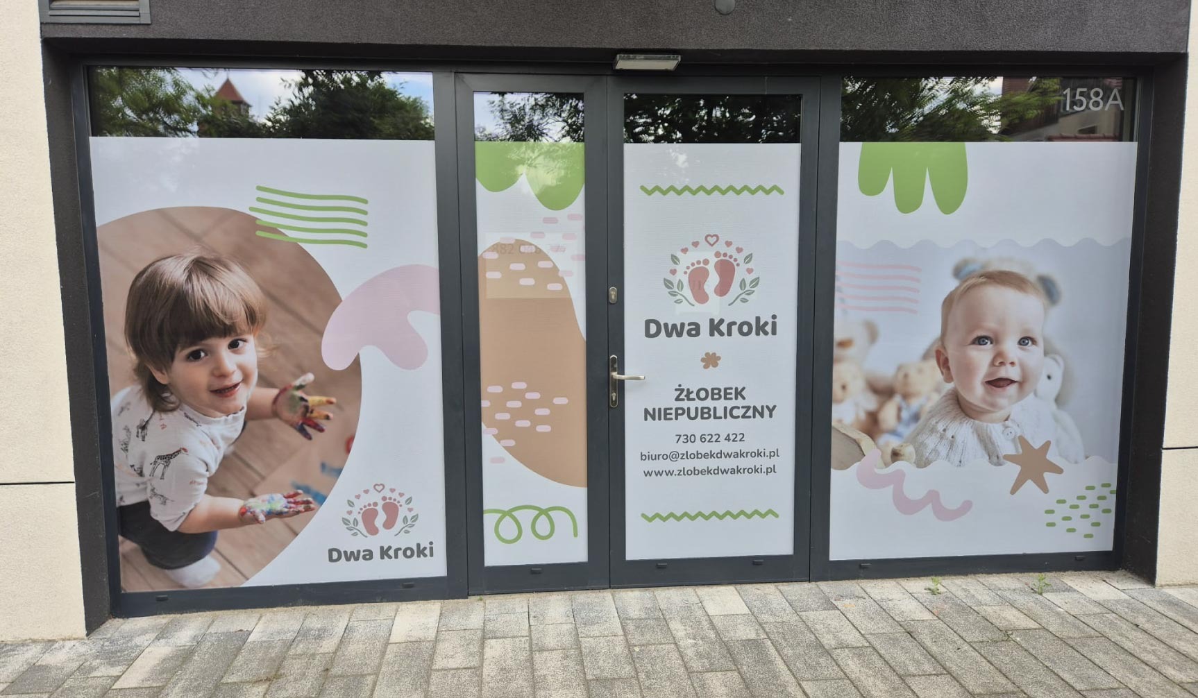 Okna żłobka 'Dwa Kroki' z naklejkami prezentującymi logo, dane kontaktowe i wizerunki dzieci, tworzące przyjazną i kolorową identyfikację wizualną placówki.