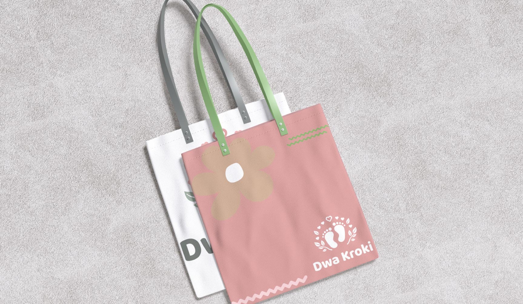 Dwie torby bawełniane z nadrukiem: biała z inicjałami i różowa z kwiatem i stopkami, leżące na szarym tle. Torby mają uchwyty w kolorze szarym i zielonym.