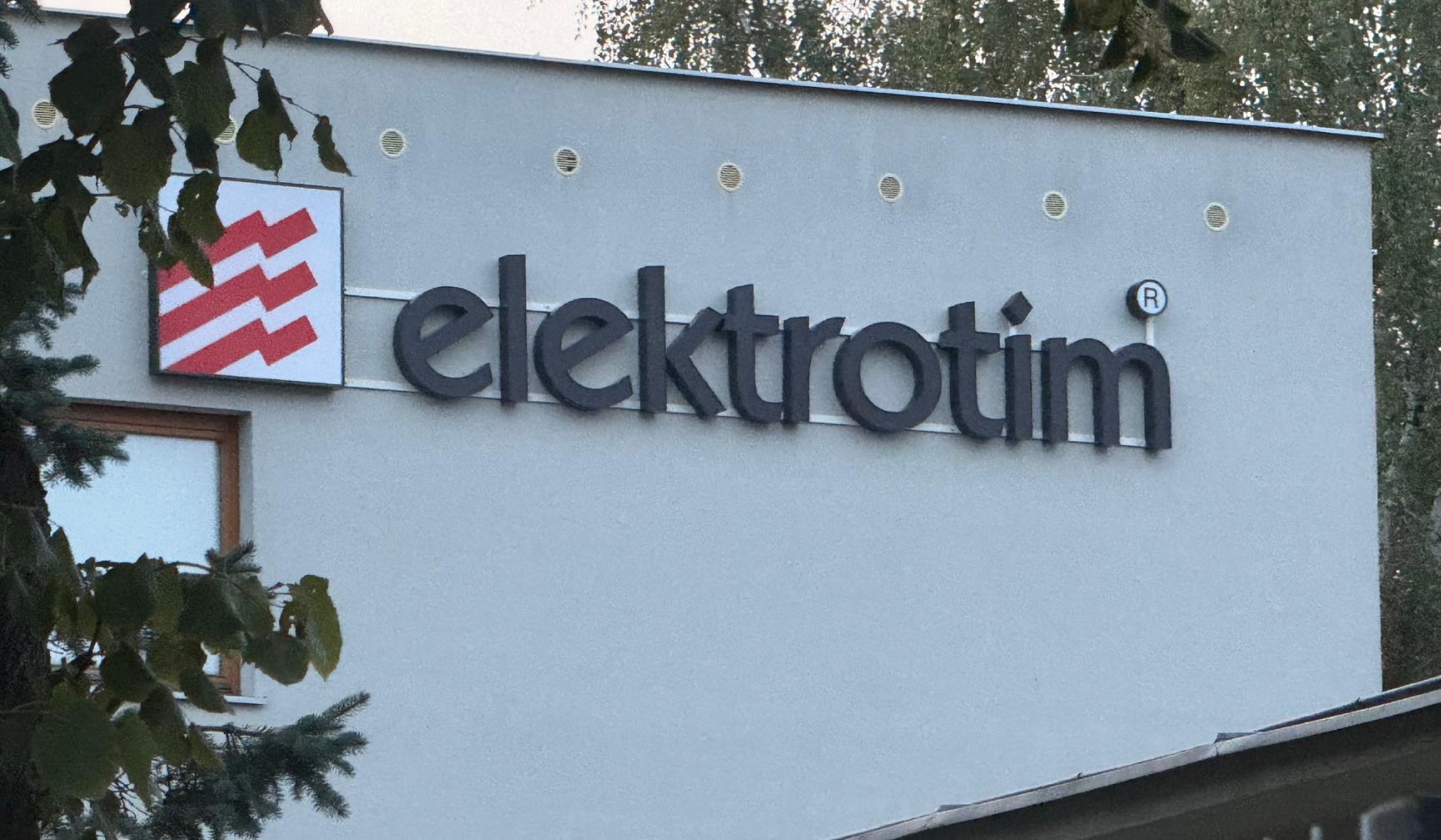 Elewacja budynku z logo i nazwą firmy 'elektrotim' w kolorze czarnym. Logo po lewej stronie, nazwa firmy po prawej. Widoczne drzewa i okno.