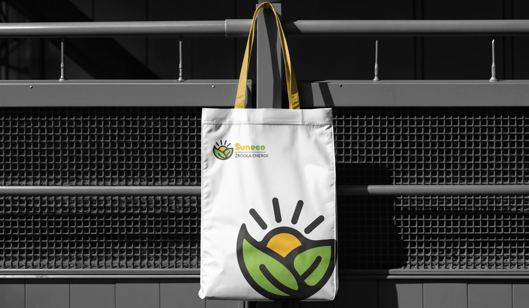 Biała torba z żółtymi uchwytami, wisząca na metalowym ogrodzeniu. Na torbie logo 'Suneeco' i grafika przedstawiająca słońce w liściach. Tło rozmyte, czarno-białe.