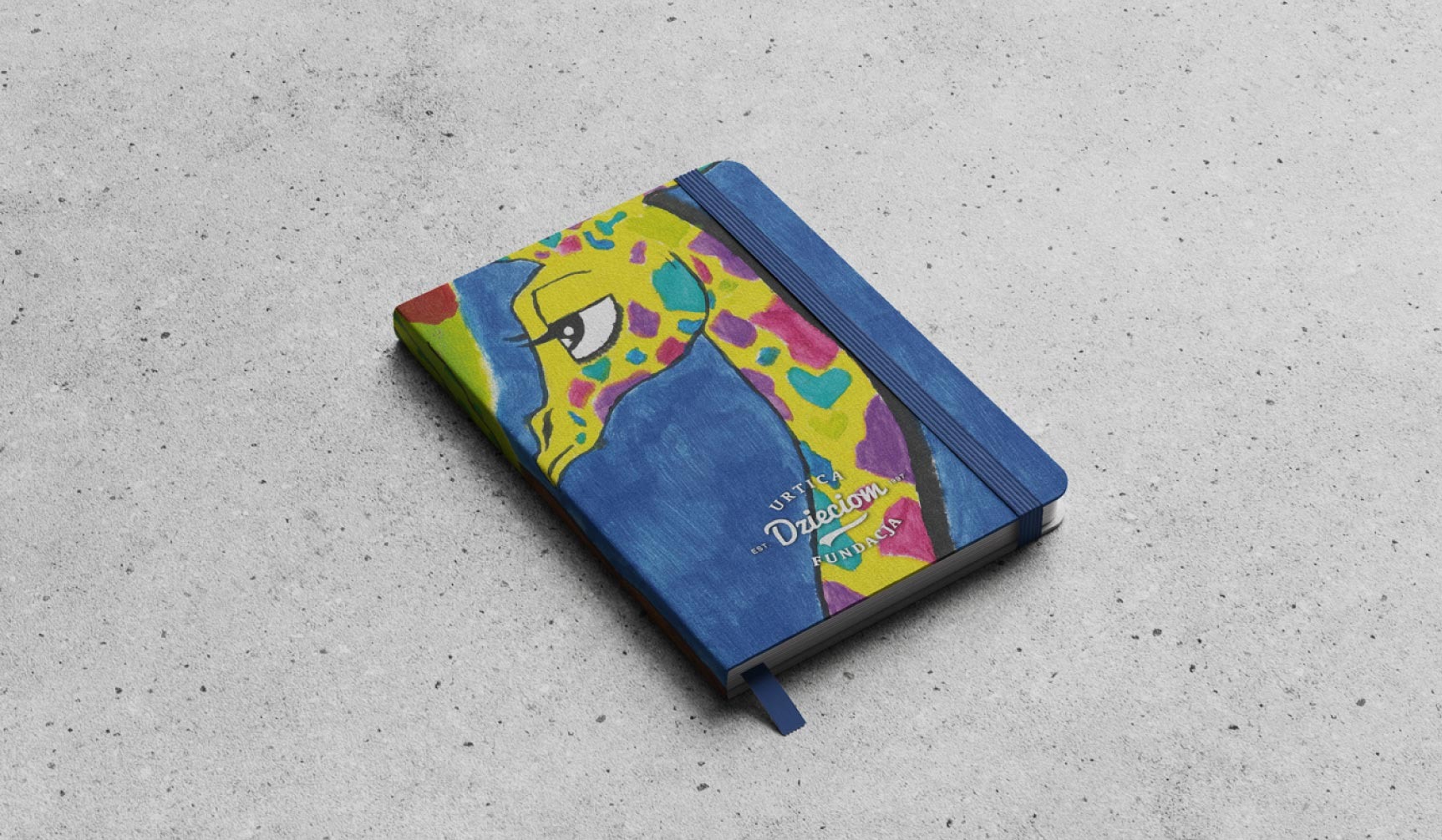 Niebieski notatnik z rysunkiem żyrafy w stylu dziecięcym, leżący na szarym tle. Na okładce widoczny napis 'Urtica Dzieciom Fundacja'.