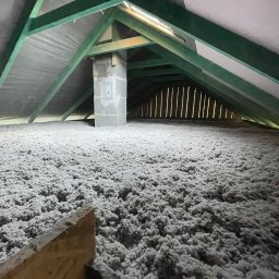 BudMar - Poddasze z widoczną warstwą izolacji z wełny mineralnej, drewniana konstrukcja dachu pomalowana na zielono, fragment komina. Widok z dołu na ocieplone poddasze.