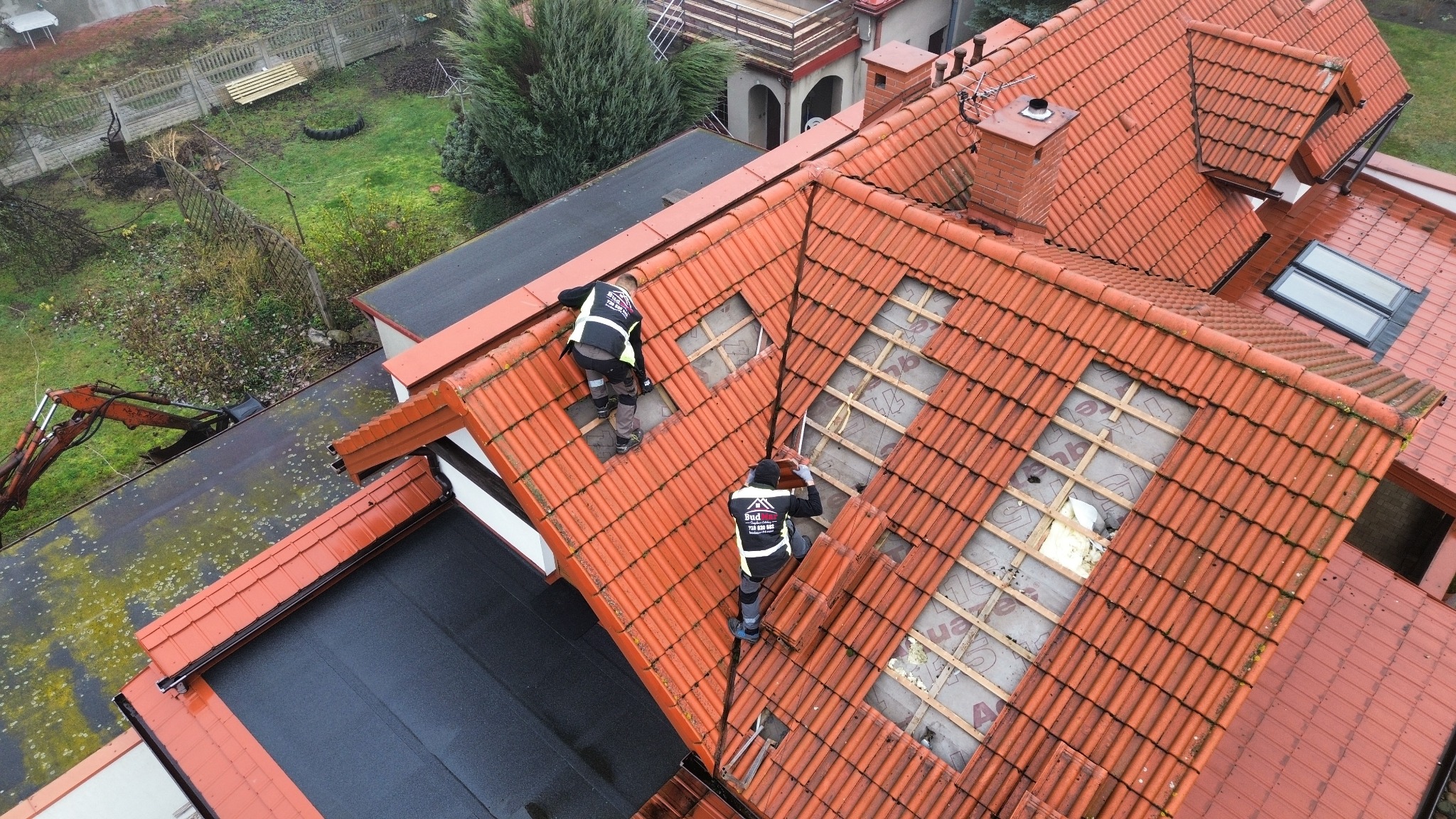 Demontaż dachówki na dachu domu z widoczną konstrukcją i izolacją. Dwóch pracowników w odblaskowych kamizelkach wykonuje prace. Widok z góry, dachówka ceramiczna.