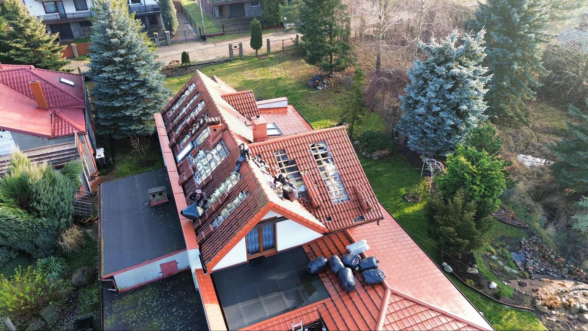 Demontaż uszkodzonej dachówki na dachu domu jednorodzinnego z widocznymi otworami i pracownikami w trakcie pracy, z workami na odpady. Ujęcie z góry.