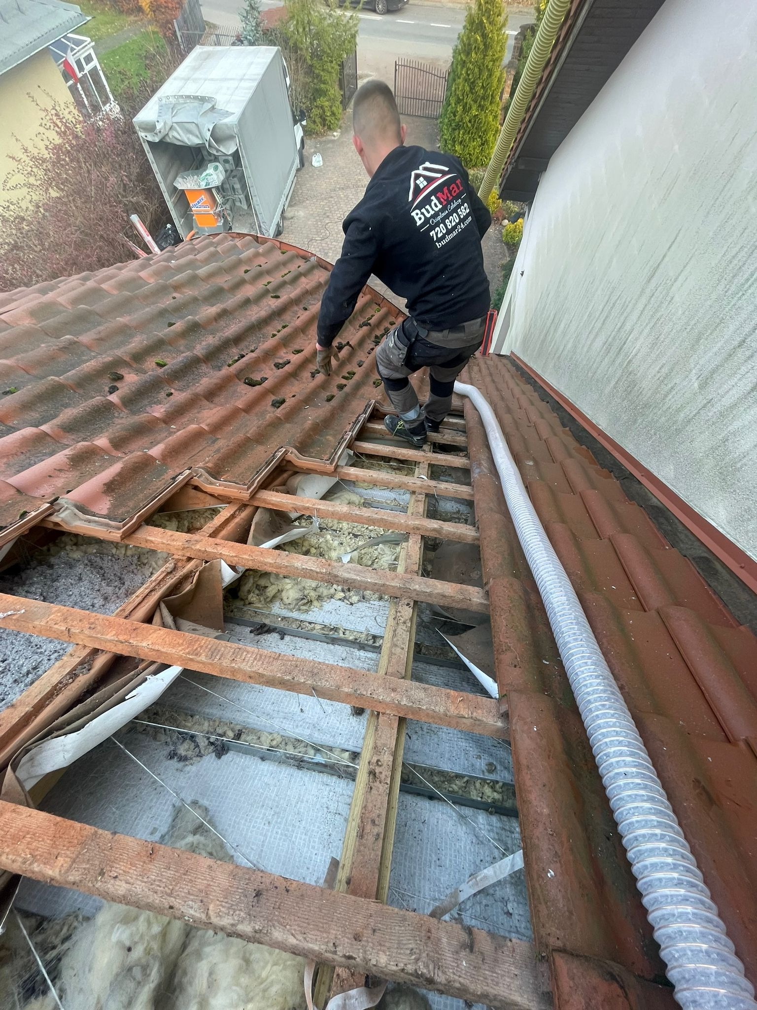 Prace ociepleniowe poddasza z widoczną konstrukcją dachu, izolacją i pracownikiem w firmowej bluzie. Na dachu widoczny wąż do wdmuchiwania izolacji.