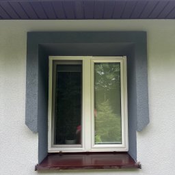DanKubconstruction - Biała ściana z oknem w szarej ramie i brązowym parapetem. Widoczna faktura tynku i fragment dachu z rynną. W oknie odbija się zieleń drzew.