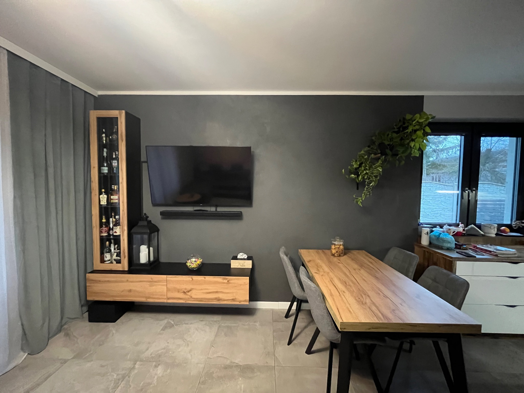 Nowoczesne wnętrze: szara ściana, drewniany stół z szarymi krzesłami, szafka RTV z barkiem, telewizor i dekoracje. Minimalistyczny design z akcentem roślinnym.