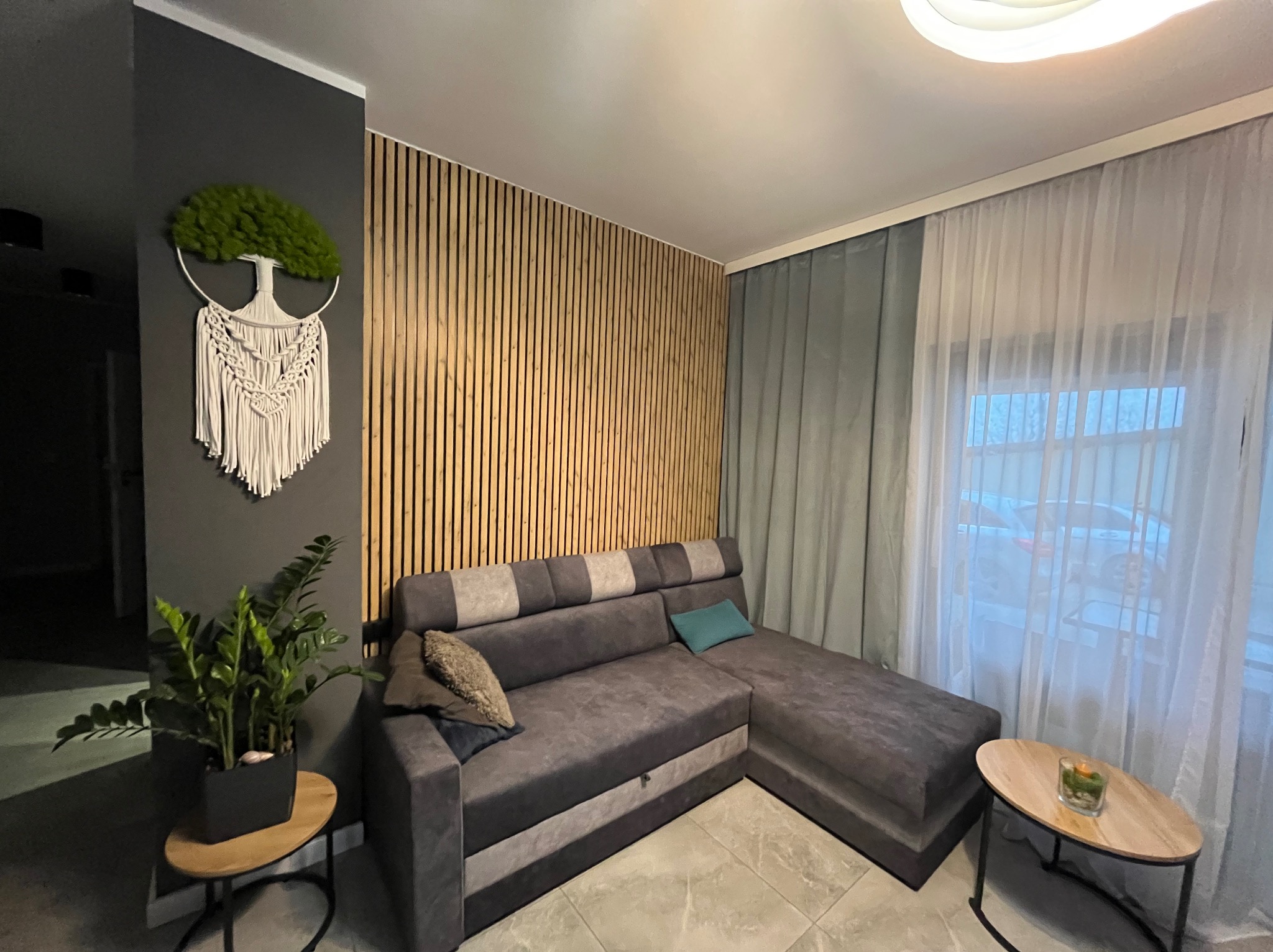 Nowoczesny salon z szarą sofą narożną, drewnianą ścianą z lameli i dekoracją ścienną w stylu makramy z mchem. Stonowana kolorystyka, minimalistyczny design.
