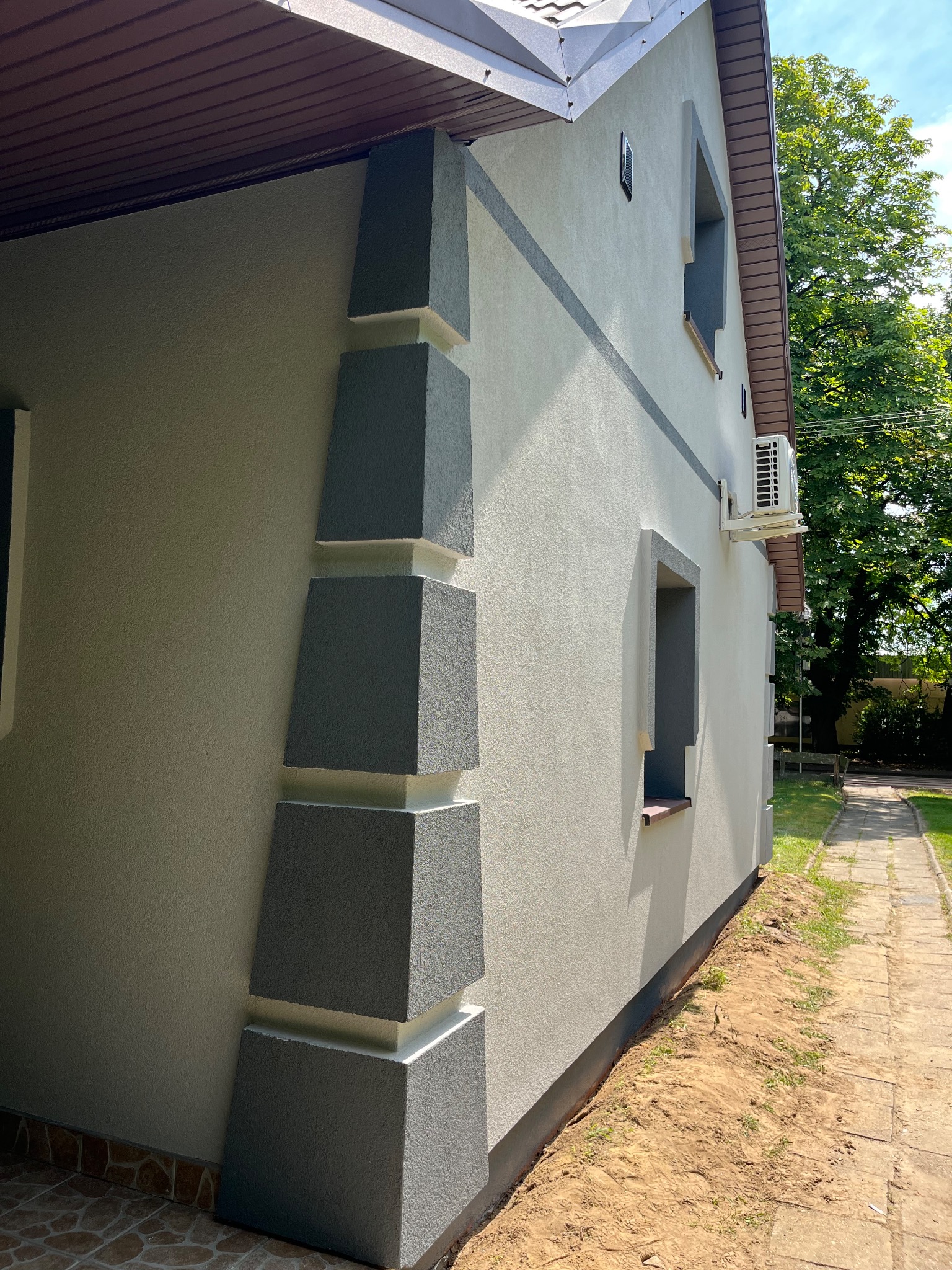 Wykończona elewacja domu z dekoracyjnymi, szarymi elementami narożnymi i listwami. Widoczny klimatyzator na ścianie i fragment chodnika.