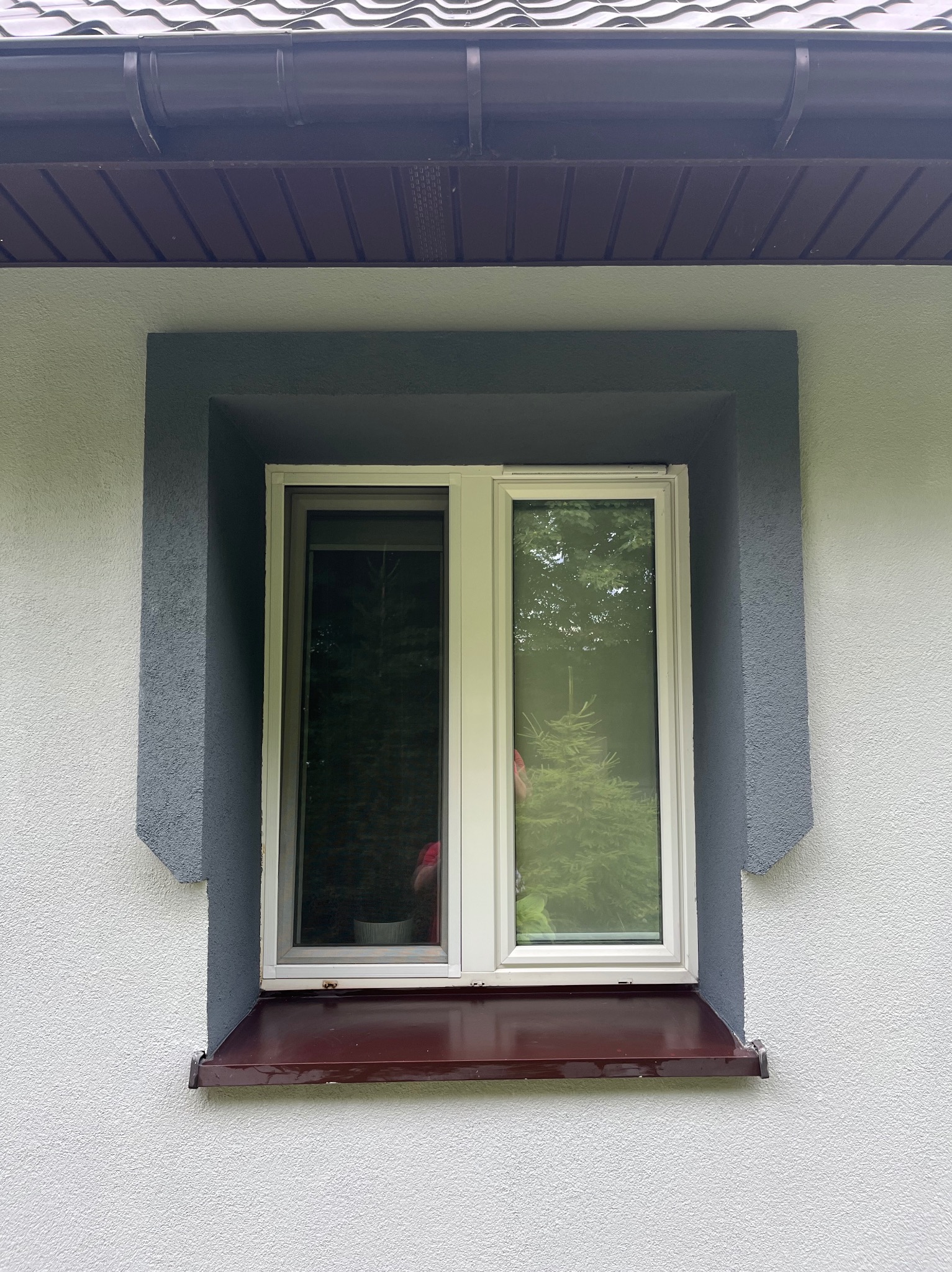 Biała ściana z oknem w szarej ramie i brązowym parapetem. Widoczna faktura tynku i fragment dachu z rynną. W oknie odbija się zieleń drzew.