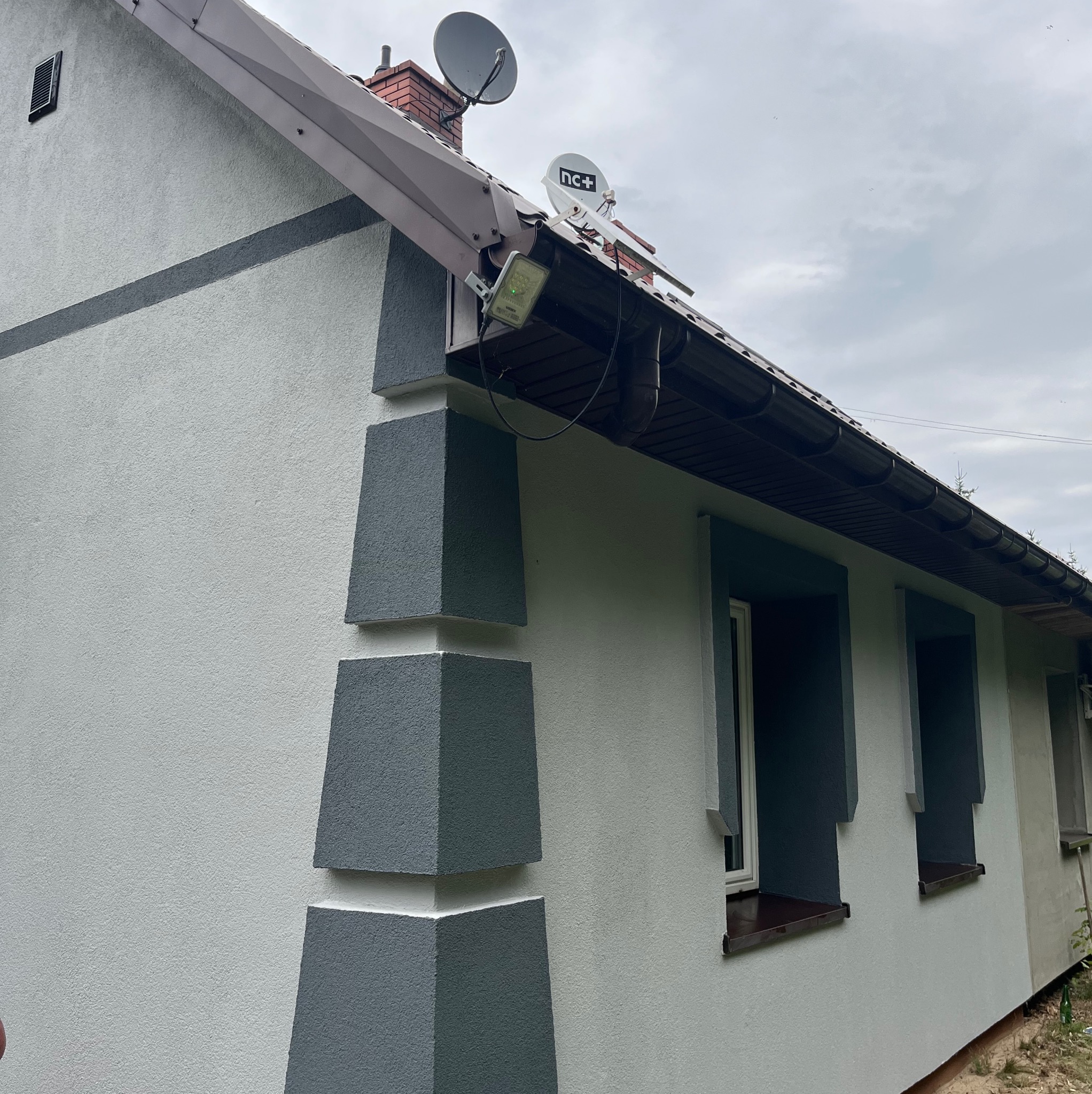Odnowiona elewacja domu jednorodzinnego z szarą, dekoracyjną strukturą wokół okien i narożników. Widoczne satelity na dachu i oświetlenie zewnętrzne.