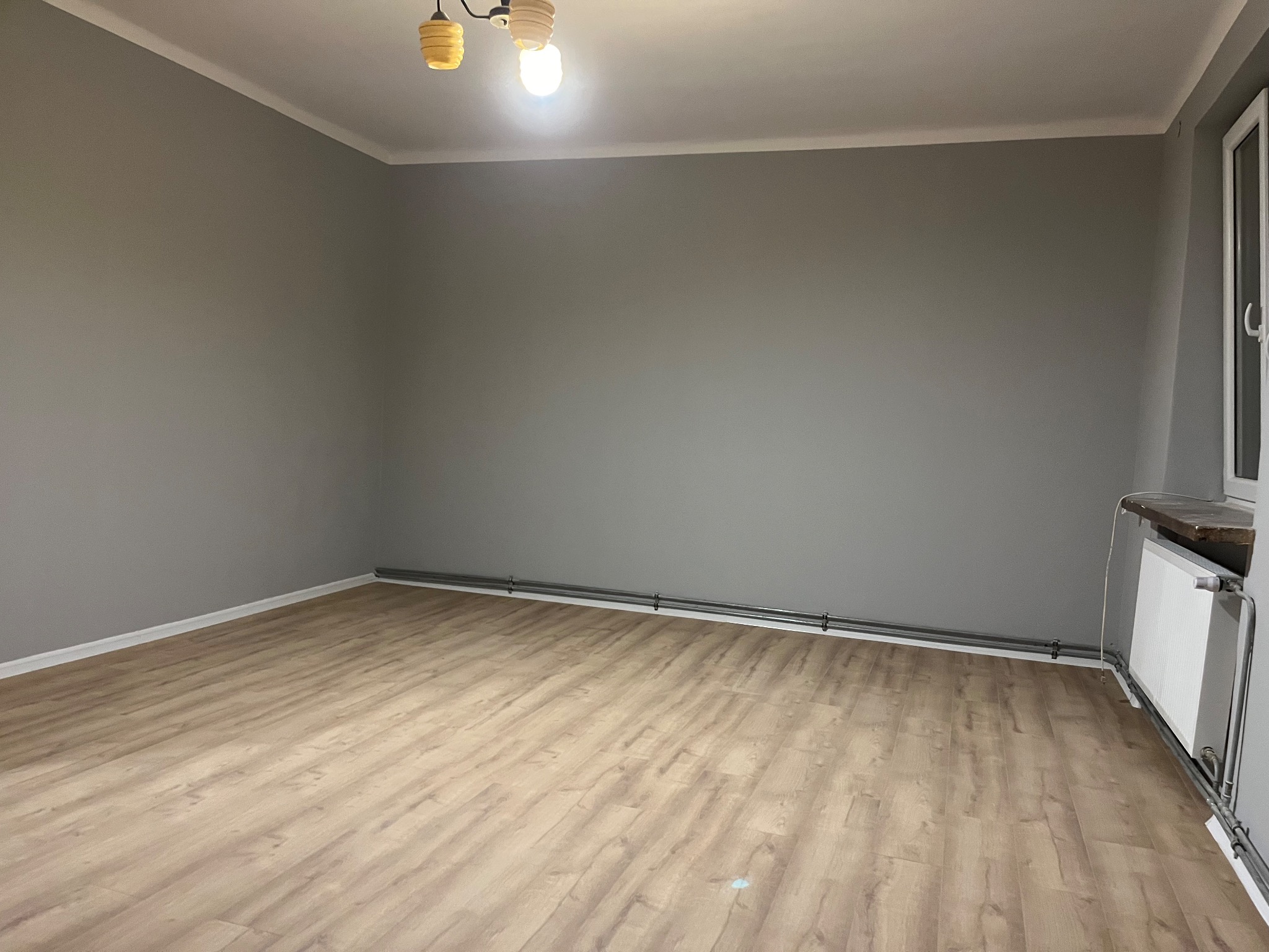 Szare ściany i panele podłogowe w pokoju po remoncie w Radomiu. Widoczny grzejnik i lampa sufitowa. Minimalistyczne wykończenie wnętrza.