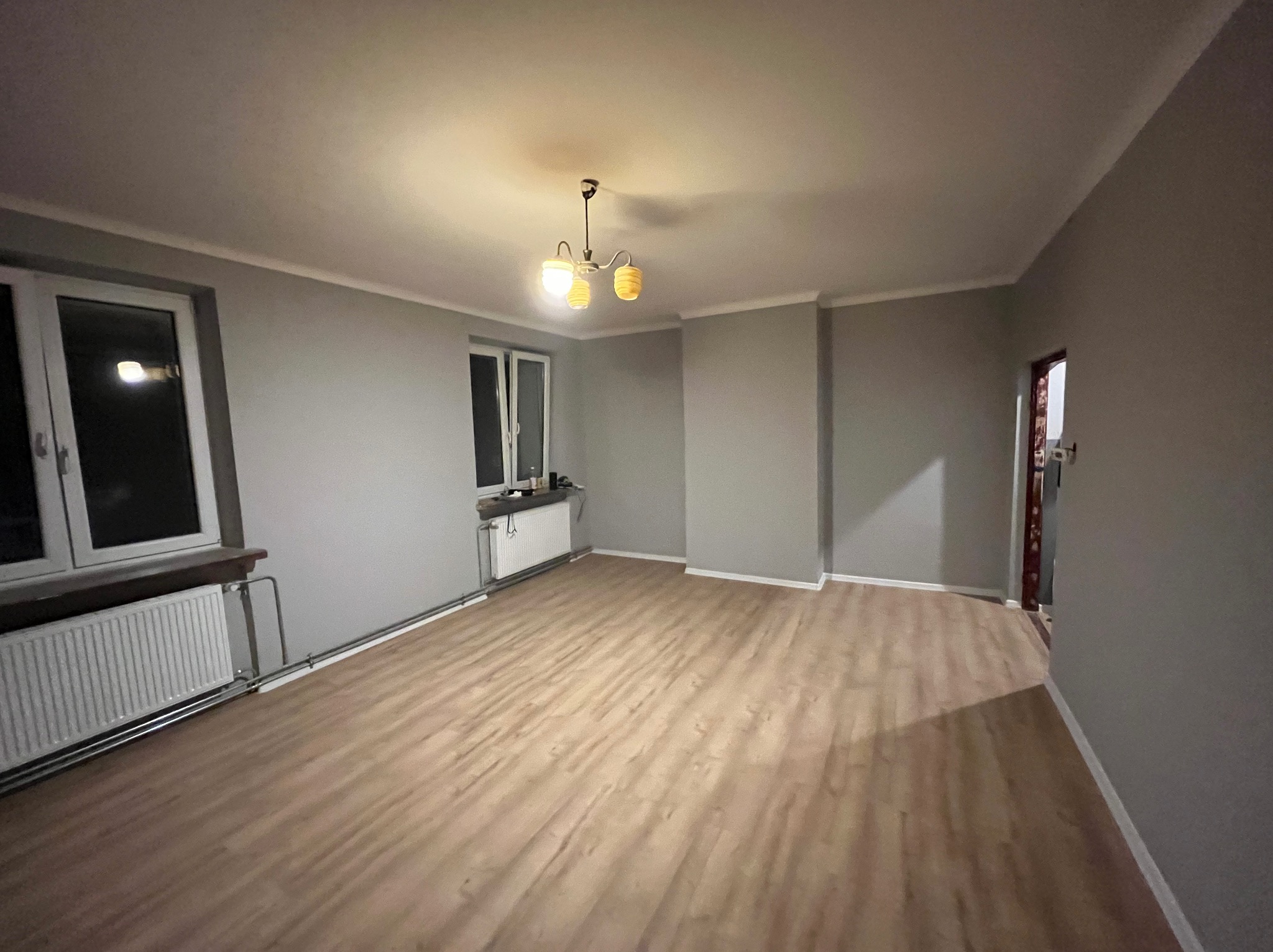 Pomalowane na szaro wnętrze pokoju z panelami podłogowymi. Widoczne okna z grzejnikami, lampa sufitowa i listwy przypodłogowe. Świeżo po remoncie.