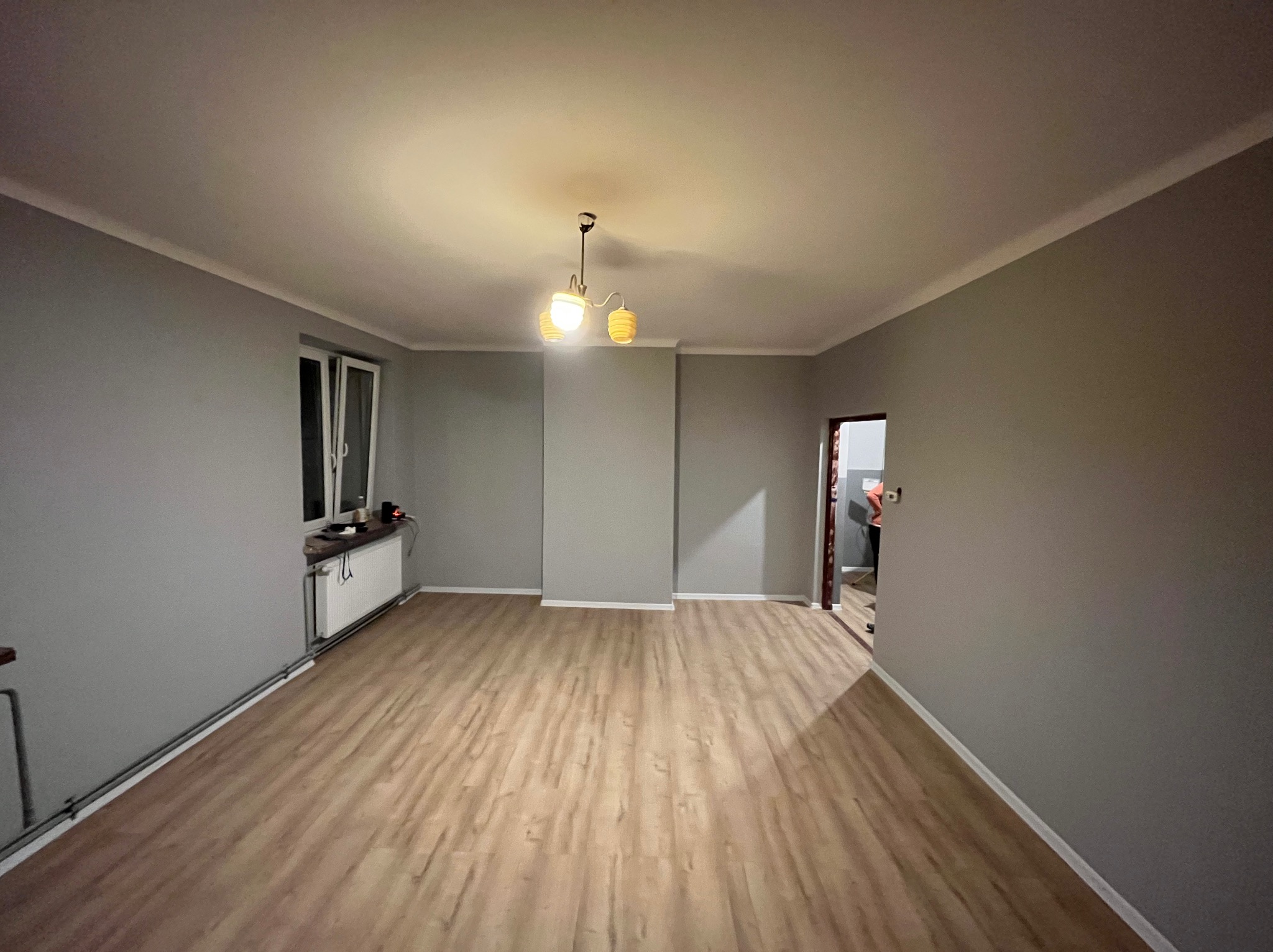 Pomalowane na szaro wnętrze pokoju z panelami podłogowymi, widoczne okno, grzejnik i lampa sufitowa. Świeżo wykończone wnętrze po remoncie.