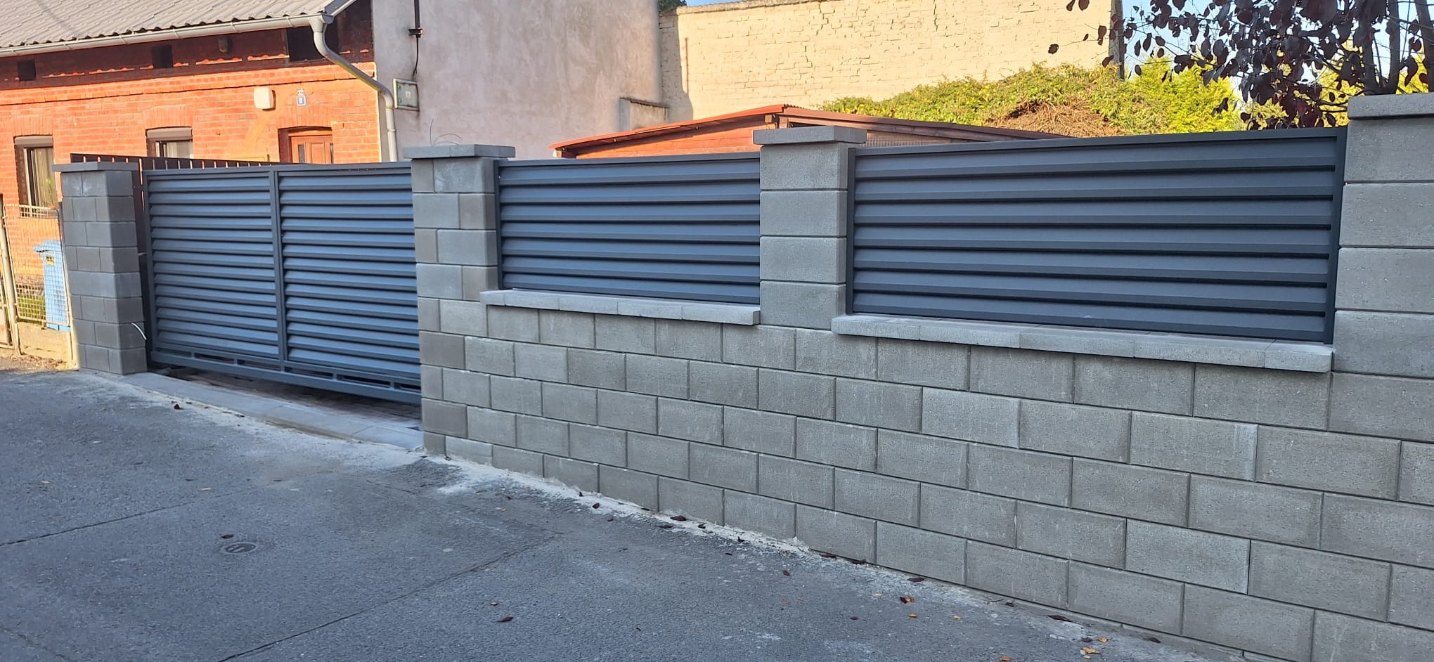 Nowoczesna, przesuwna brama wjazdowa z szarymi, poziomymi panelami, osadzona na murze z szarego betonu architektonicznego. Widok z zewnątrz posesji, w tle budynki.