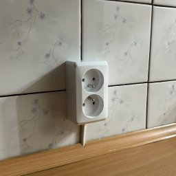 Global Alpha Szymon Krywult - Białe, podwójne gniazdko elektryczne wystające ze ściany wyłożonej kwadratowymi płytkami z delikatnym, błękitnym wzorem, nad drewnianym blatem.