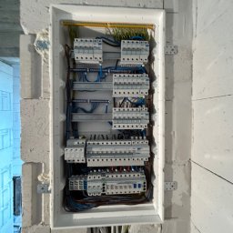 Global Alpha Szymon Krywult - Rozdzielnia elektryczna z zabezpieczeniami, zamontowana w surowej ścianie z betonu komórkowego. Widoczne przewody i elementy instalacji.