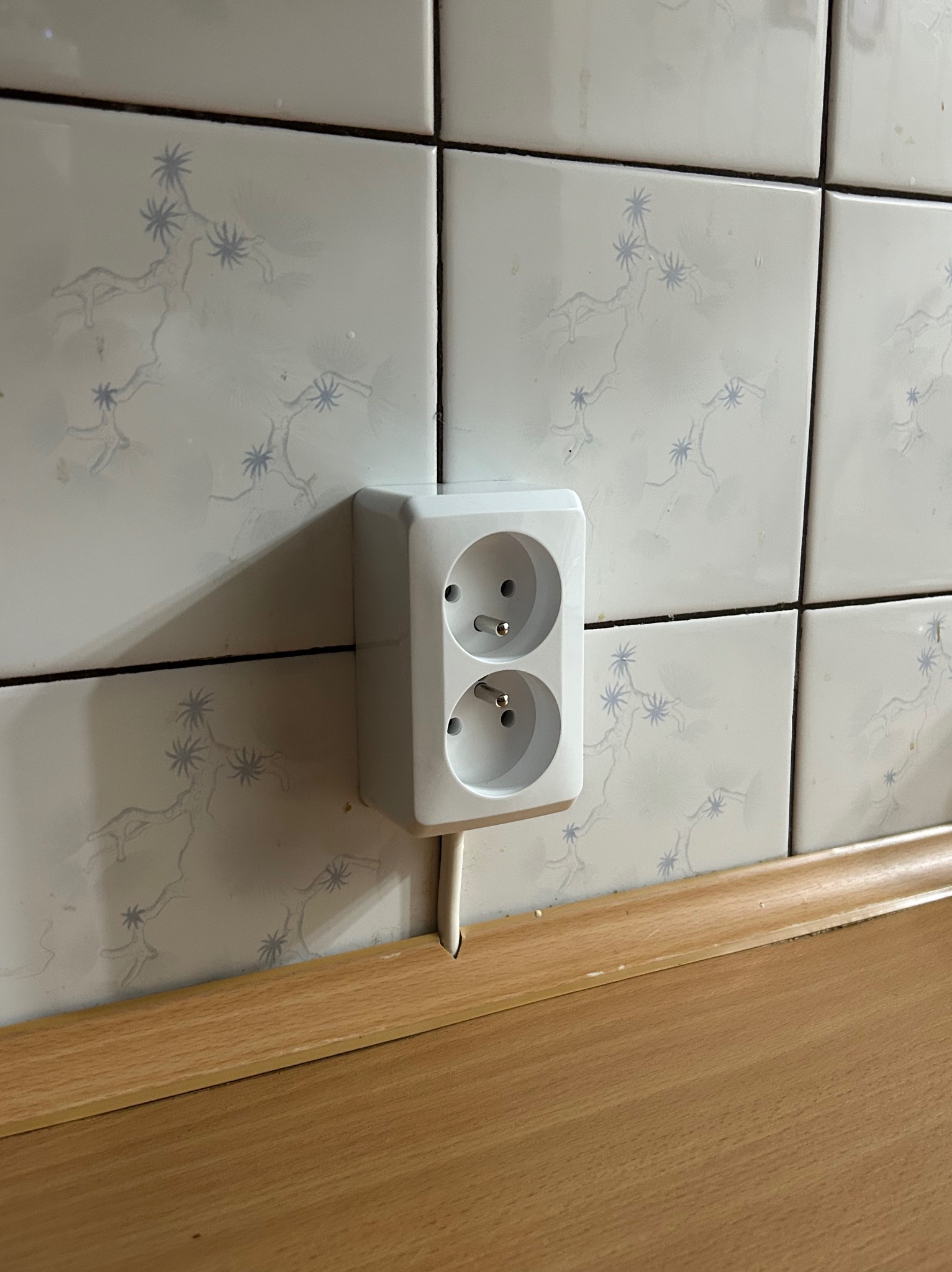 Białe, podwójne gniazdko elektryczne wystające ze ściany wyłożonej kwadratowymi płytkami z delikatnym, błękitnym wzorem, nad drewnianym blatem.