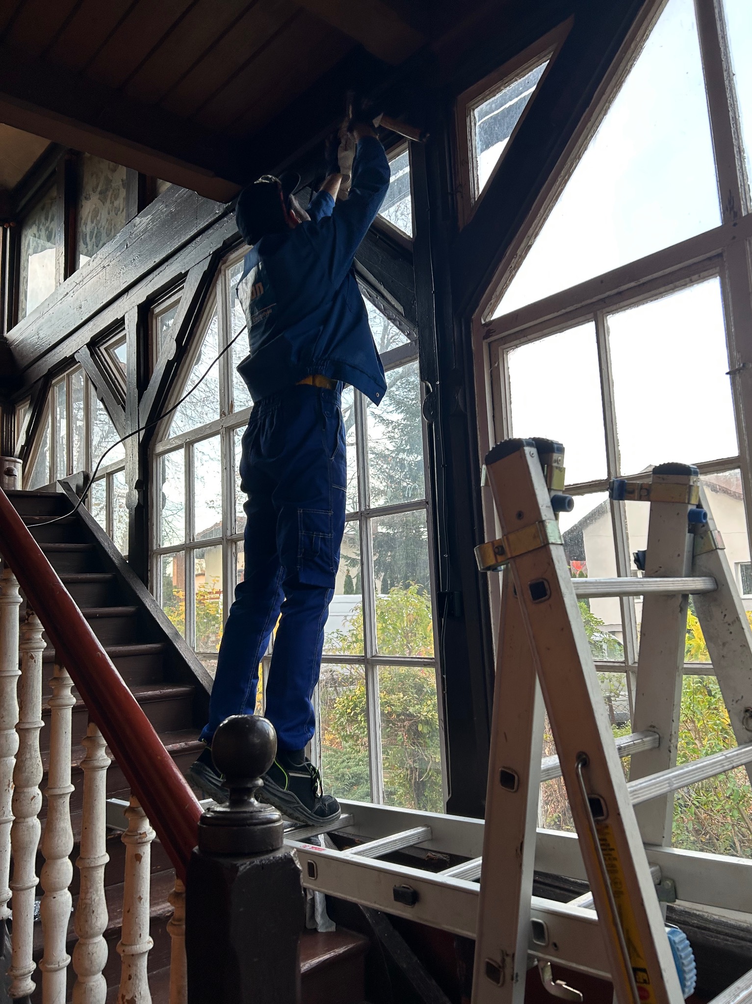 Elektryk w niebieskim kombinezonie, stojąc na schodach, montuje oświetlenie w zabytkowej werandzie z dużymi oknami. Widoczna drabina i narzędzia.