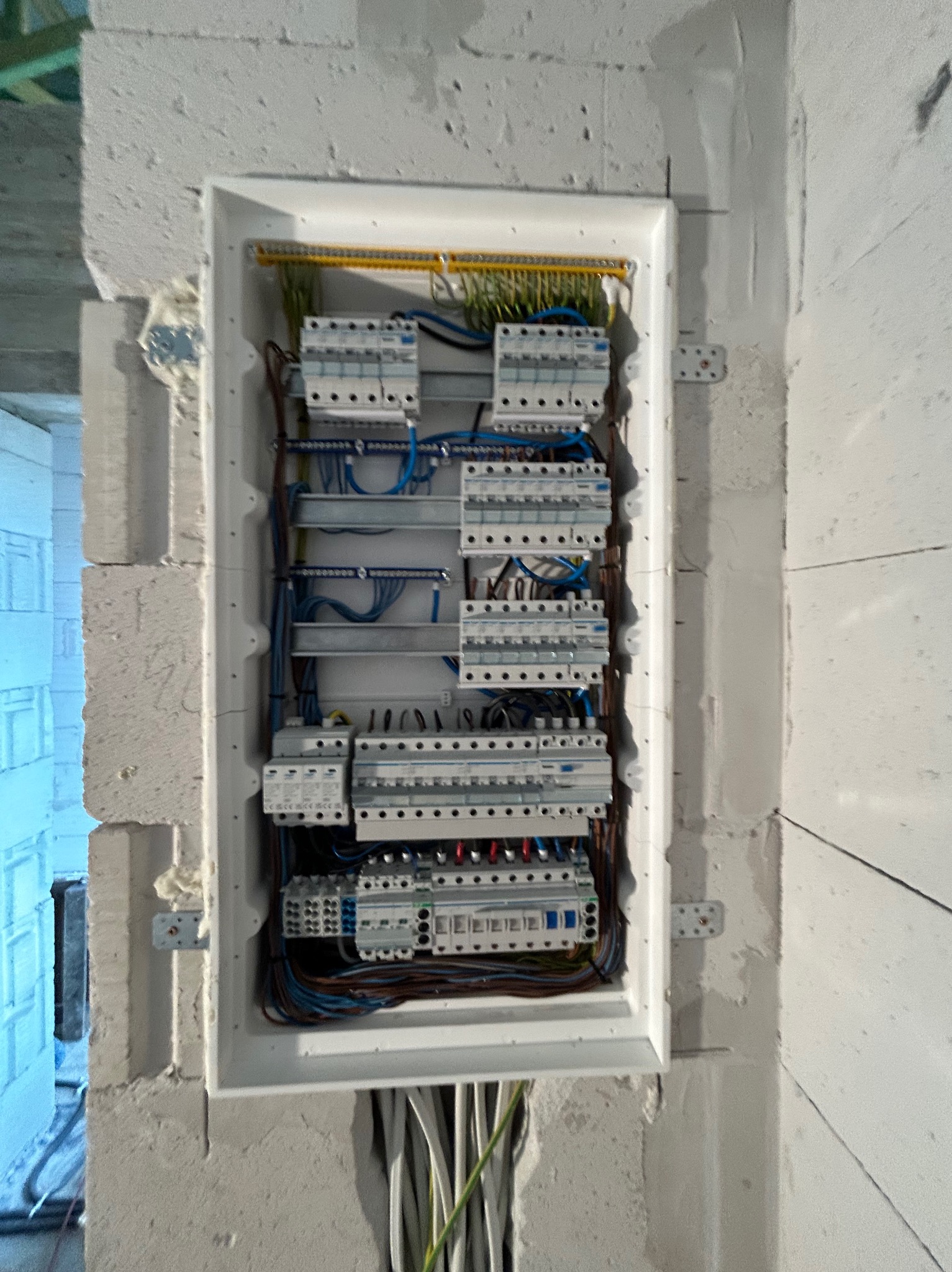 Rozdzielnia elektryczna z zabezpieczeniami, zamontowana w surowej ścianie z betonu komórkowego. Widoczne przewody i elementy instalacji.