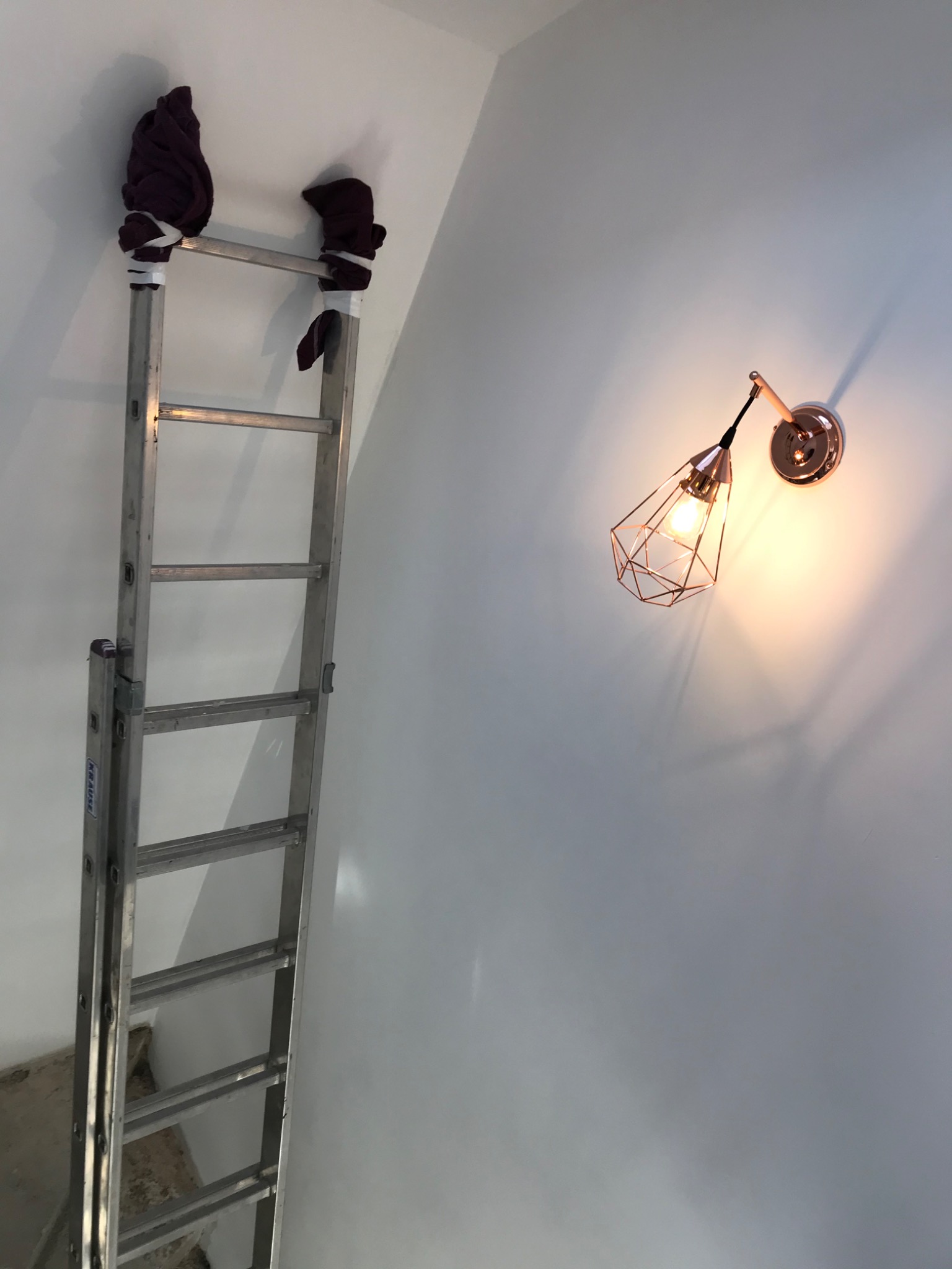 Zamontowany kinkiet w stylu industrialnym na białej ścianie, obok stoi drabina z zabezpieczeniami na górze. Widok z dołu, oświetlenie ciepłe.