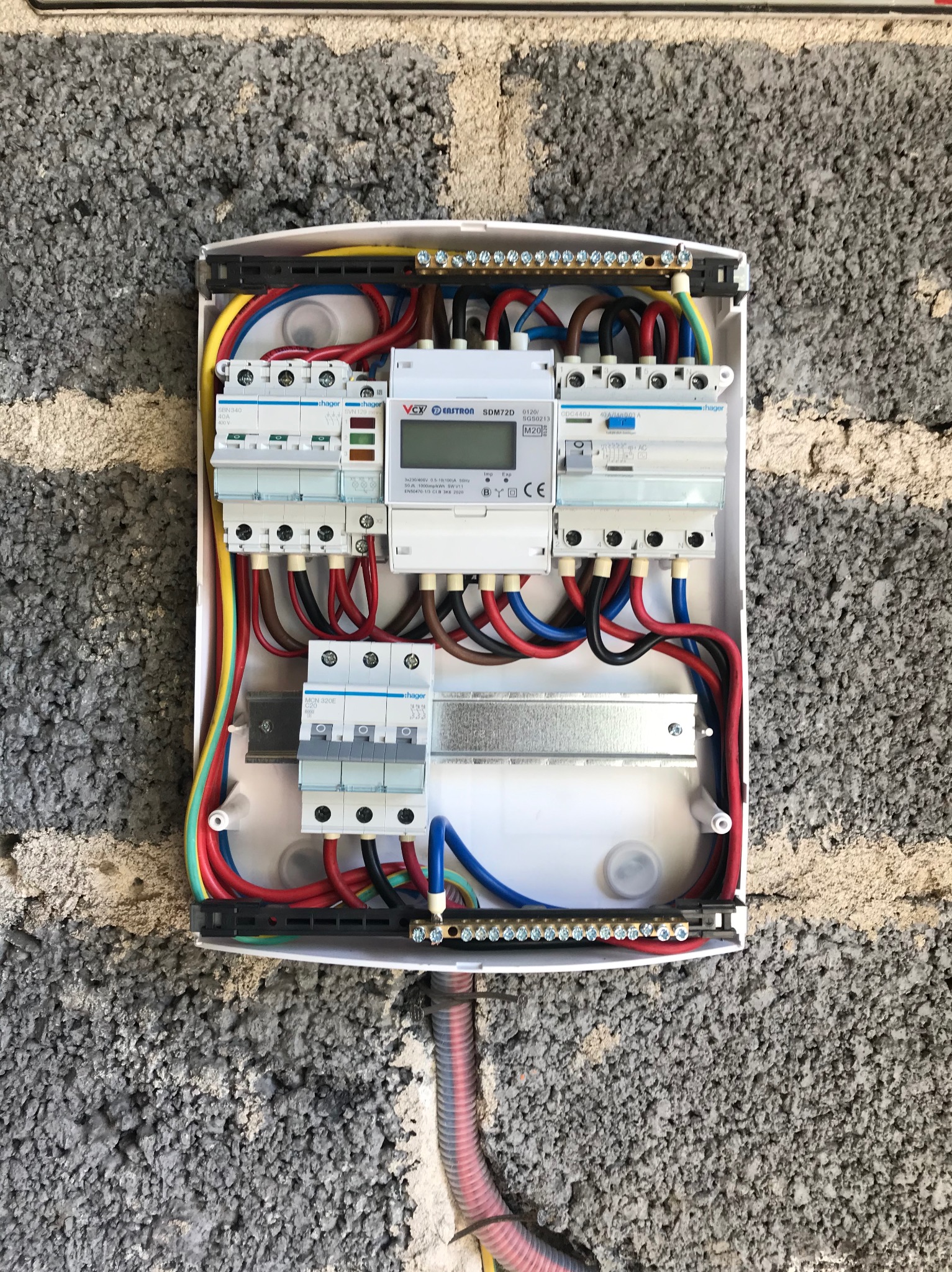 Rozdzielnica elektryczna Hager z zainstalowanymi bezpiecznikami i licznikiem, zamontowana na szarej ścianie z betonu komórkowego. Widoczne przewody w różnych kolorach.