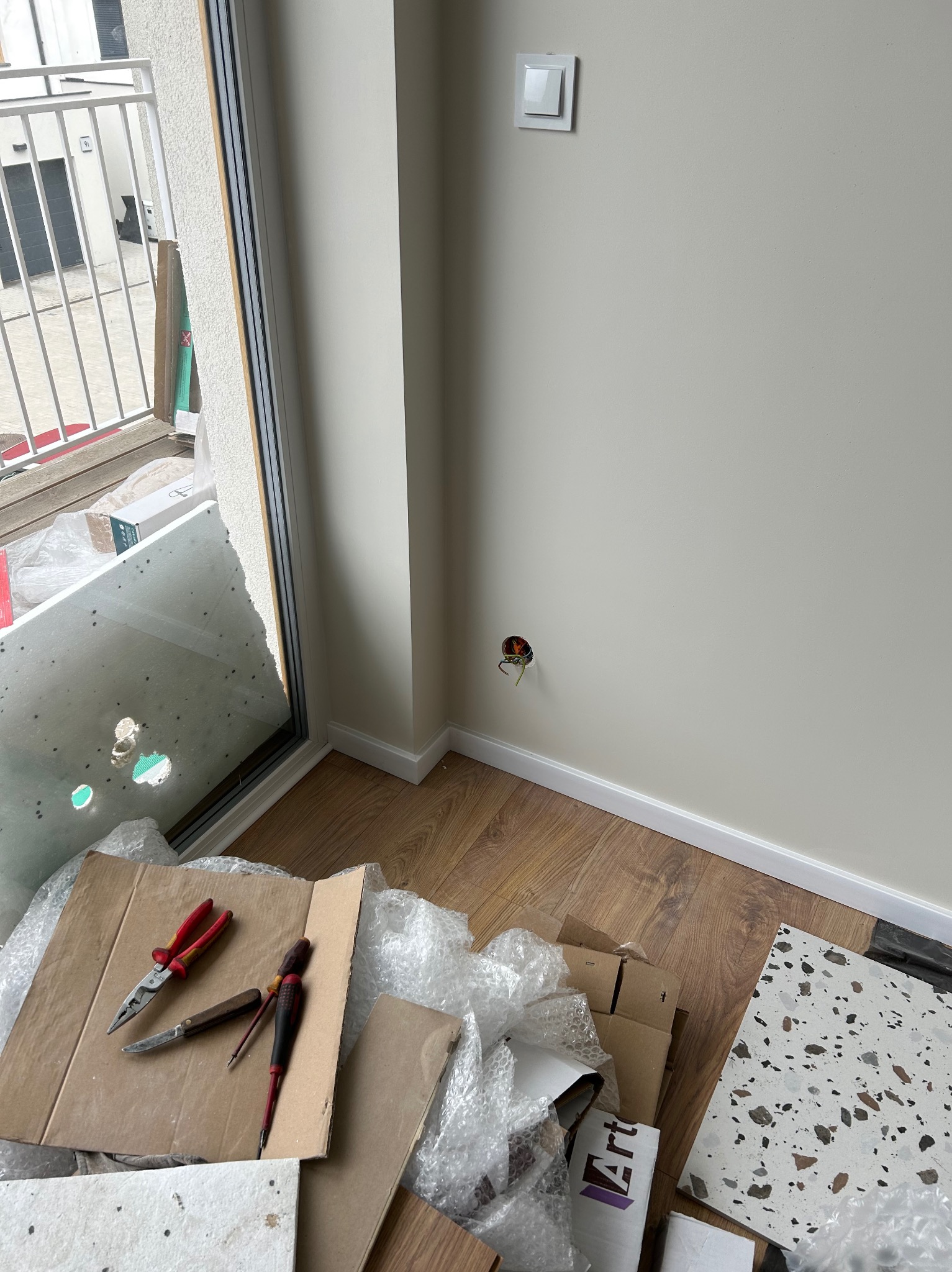 Gniazdko elektryczne w ścianie, obok narzędzia i materiały budowlane. Widoczne kable, włącznik światła, szczypce, śrubokręty i płytki podłogowe.